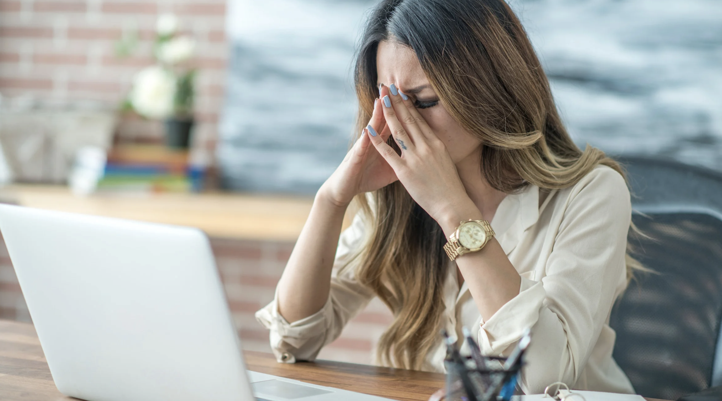 Stressed woman with fingers pressing on her eyebrows as she sits at a desk with her eyes closed.